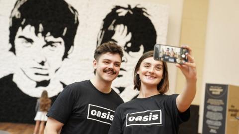 A male and female Oasis fan take a selfie in front of a monochrome portrait of Oasis bandmates Noel and Liam Gallagher constructed entirely from bucket hats on display at the shopping centre in Cardiff, ahead of the band's first comeback tour gig