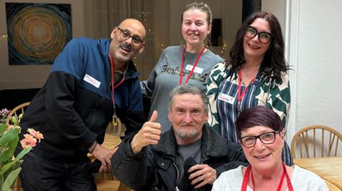 Five people, in a room, all looking at the camera. A woman, and a man are sitting down, the man has on a dark coat, top and trouser and has a thumb up. He has a plate of food and a cup by him on a table, with flowers on. The woman next to him has purple hair and is wearing a white top with a red apron on. Three other people are standing behind, with lanyards on all smiling. 