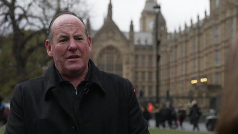 Alan Smith outside Parliament, wearing a black coat and short hair.