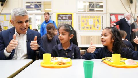 Mayor of London Sadiq Khan sitting at a lunch table with two school children, doing a thumbs up. He is wearing a dark blue suit and white shirt. The two female pupils have yellow plastic trays with food and a plastic cup in front of them on the table. One of the children is also doing a thumbs up.  