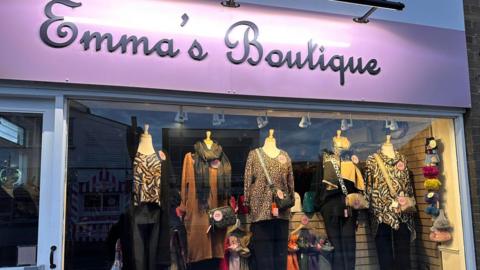 A shop front full of women's clothing. The sign at the top in black text on a pink background reads Emma's Boutique. 