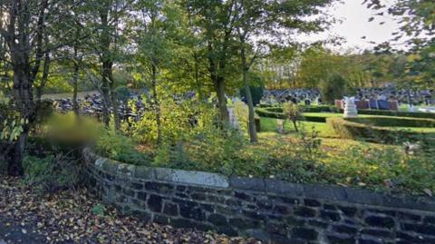 A cemetery showing burial plots in the background behind a stone wall and beyond a row of trees on a sunny day.