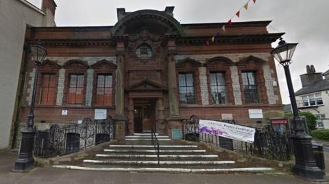 Kendal Library in Coniston. The stone building has two columns near the entrance. Stone steps with a metal handrail leads to the front doors.