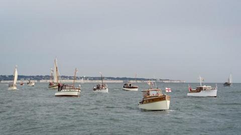 Multiple ships dating back from the 1940s sailing in the sea. Some of the ships are flying St George's flags. The English coast is sat in the background.