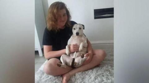 Ellis Cox, with shoulder length brown hair, sits smiling cross-legged on a fluffy rug with his white dog sitting on his lap. 