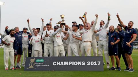 Essex County Cricket Club players celebrate winning the County Championship in 2017 by punching the air and spraying champagne. They are wearing their whites on the field, although a handful of them are wearing navy.