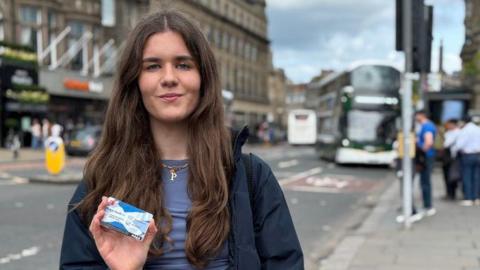 Poppy Fairbairn holds the National Entitlement Card towards the camera with a blurred Lothian Bus in the background.