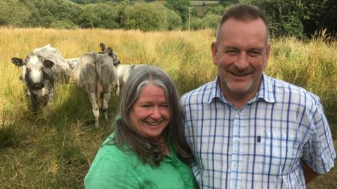 Emma and Andrew arm in arm in one of their fields with three of their Albion cattle laying behind them. Emma is wearing a bright green shirt, Andrew a white and blue chequered shirt, both smiling. 

The cattle appear a greyish blue and white mottled colour, one of the cows is facing the camera.