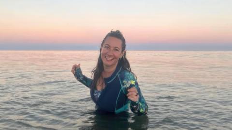 A woman in a wet suit standing on the sea, the sun setting behind her 