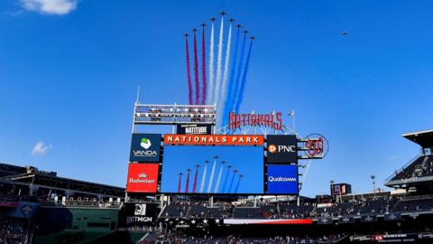 The red arrows planes have red, white and blue colour trails behind them as they fly in formation over a sports stadium, there is a large screen with advertisements in the centre of the photo and crowds sat in the stands.