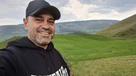 A man in his early 40s, wearing a baseball cap and smiling into the camera. He has grey stubble and is standing against the backdrop of some green hills.