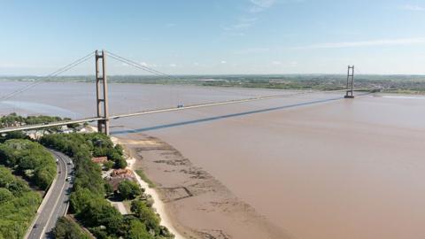 Aerial view of the Humber Bridge from the north bank of the Humber Estuary showing the bridge spanning the muddy water with vehicles crossing and traffic on the A63 road by the side of the bridge