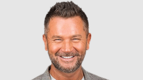A man with short spiky brown hair is smiling at the camera in a company promo shot. He is wearing a grey jacket and standing in front of a pale background.