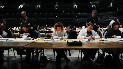 Row of people sit at desk in large arena. They're sitting with their heads looking down at the table as they count ballots