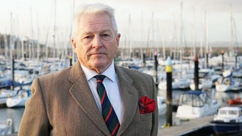 Col Tim Collins, wearing a suit, looks into the camera on Bangor marina with boats behind him