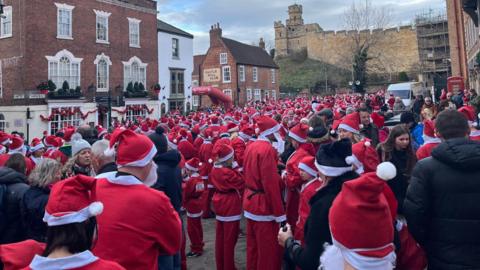 Hundreds of people dressed up in the red and white outfit of Santa Claus, some wearing white beards, stand shoulder to shoulder in a large group in a cobbled square beneath the imposing walls of a medieval castle. To the left, Georgian-style buildings can be seen, including one advertised as the Castle Hill Club.