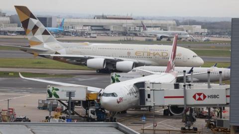 Various passenger jets unloading or taxiing at Heathrow airport with a Virgin plane in the foreground and an Emirates plane behind it