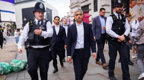 Sadiq Khan, a middle-aged man wearing a suit, walking on a busy shopping street with male police officers walking either side of him.