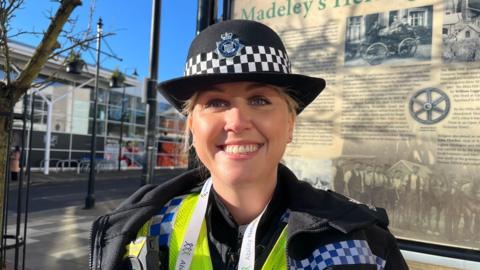 Sgt Webb pictured looking into the camera and smiling. She's wearing police uniform; a black jacket over the top of a yellow hi-vis vest, with another black top underneath that. She's wearing a black police bowler hat with the West Mercia Police badge on it. Behind her is a beige sign, with the top of it just visible and reading 'Madeley.' The sign celebrates the area's heritage with black and white photographs of the streets in the past. Over her right shoulder is a big Tesco. The sky is clear and bright blue with lots of sunshine. 