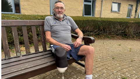 A man is sitting on a wooden bench with a green hedge behind him and a building with blue windows. He is wearing a grey shirt and has a synthetic leg which is largely black but has a white section on the end