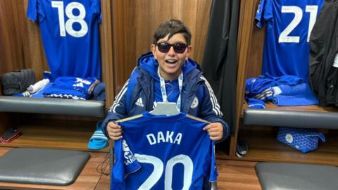 Teddy, wearing lots of Leicester City clothing and holding Patson Daka's shirt