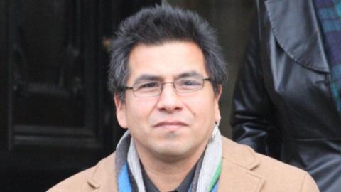 A 47-year-old man with spiky hair and glasses wearing a tan-coloured coat, a scarf and First Nations insignia