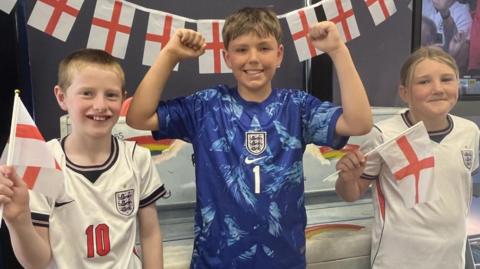Three pupils stand smiling the the camera. They are wearing England football kits. 