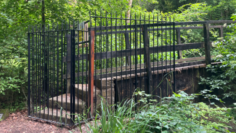 A large black metal gate with spiked railing blocks steps leading to a bridge. The surrounding area is green and wooded.