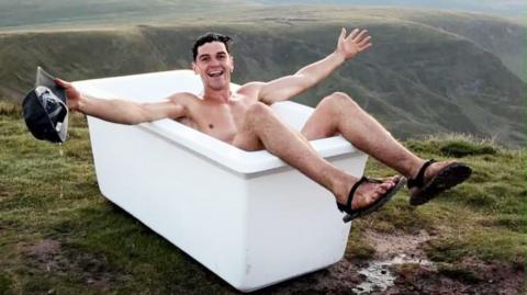 A man is bathing in a bathtub posing for a camera while on a mountain summit. He has short dark hair, holds a black cap in one hand, and is wearing flip flops.