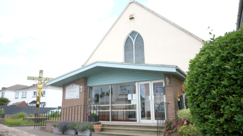 A large cross with a gold coloured image of Christ is outside a church with a brick porch area and a white sloping upper storey. A large stained glass window is in the middle of the wall.