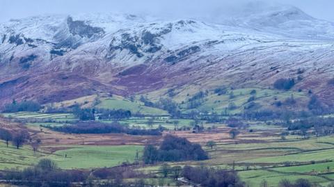 Threlkeld in the Lake District. Landscape shot of mountains which have a smattering of snow. Below the mountains are green fields with lots of trees in patches. The sky is grey. 