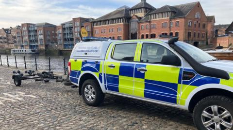 A white, blue and yellow police vehicle with the words "regional marine and underwater search unit" written on the side. There is a trailer attached to the back of the vehicle, which is parked next to a stretch of river. In the background, there are buildings overlooking the water.