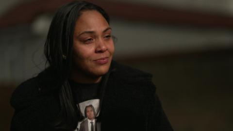 Katherine Johnson, wearing a black coat and a T-shirt with her son Kamari's photo on it. She has long, dark hair.  