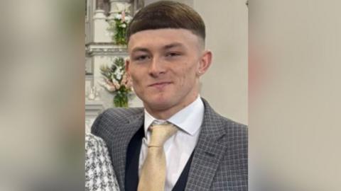 A man in a grey chequered suit, white shirt and gold tie looks at the camera. Behind him are flowers on the wall. 