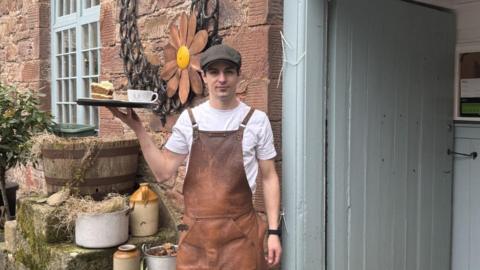 Shane Perry pictured holding a tray with a cake and drink on in front of the cafe entrance.