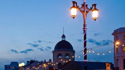 Worthing Pier at dusk. A street lamp is illuminated with a string of lights underneath. The sky is fading with some blue clouds scattered. The top of the pier is visible at the bottom of the photo. 