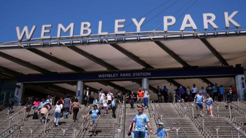 Football fans walk down stairs from Wembley Park station towards Wembley Stadium. It is clearly  sunny day. The fans are most wearing sky blue Coventry City football shirts.