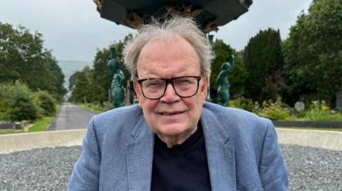 An elderly man with grey hair and black square glasses and a grey blazer on. He is standing in front of some grey stones and blue statues and what looks like a public garden with trees and grass. 