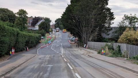 Single carriageway road with numerous cones along its edge and roadworks vehicles in the distance.
