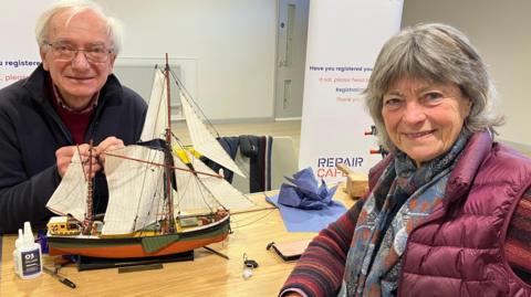 A man and a woman are sat across from one another at a table.  Both are looking to the camera.  The woman in the foreground is wearing burgundy coloured clothing with a scarf around her neck. The man in the background has glasses and is wearing a blue fleece.  Between them on the table is a model of a Thames Wherry, a boat from the 1920's with two masts and 5 sails.  It's rigging is damaged and the man is trying to repair it. 