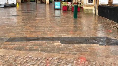 A long tarmac strip runs over some brick paving in front of a shopping centre entrance.