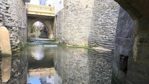 The River Lim viewed from beneath a stone bridge. The view is far below street level and the river is running through a deep channel with high stone walls either side.