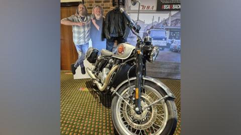 The BSA Gold Star pictured at The National Motorcycle Museum. It has a silvery-grey, chrome and black colour scheme. Behind it is a carboard cut-out of the Hairy Bikers and a leather jacket.