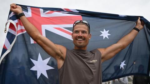 British ultra endurance runner William Goodge celebrates, holding an Australian flag, after crossing the finish line at Bondi Beach