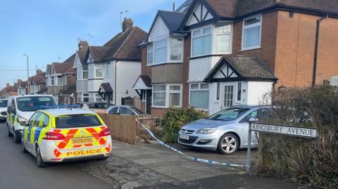 A Google Streetview look at Kingsbury Avenue. Blow's Downs are on the horizon and there's semi-detached houses on either side of the road which runs down the middle of the shot. Cars are parked half on the road and pavement