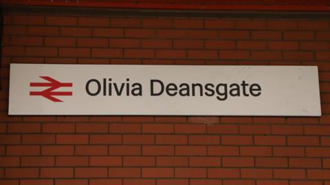 A train station sign reading 'Olivia Deansgate' hangs on a red brick station wall