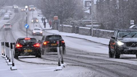 Several cars with their lights on are photographed driving on a snow-covered street.