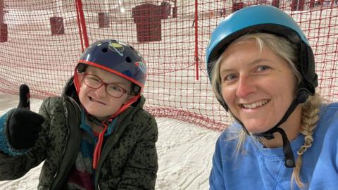 Hugo and Claire Reece on a ski slope. He is on the left and wearing glasses,  a black helmet and dark fleece and has his right thumb up while smiling. She is on the left wearing a light blue helmet over plaited blonde hair and a blue sweatshirt and also smiling. They are sitting on snow and behind them is a red net. 