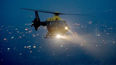 An NPAS helicopter is flying at night above a city with lit buildings. It has its lights on and along its side reads 'Police' and its aircraft number is on its tail, not quite legible. 