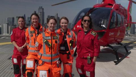 Group photo of the five female crew members on top of a building with the red helicopter behind them and a London skyline in the background